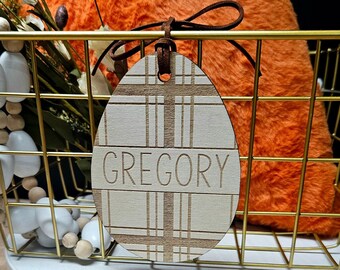May include: A wooden egg-shaped ornament with a plaid pattern and the name "GREGORY" engraved on it. The ornament is hanging from a brown ribbon and is in front of a gold wire basket.