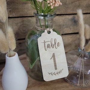 May include: A wooden table number tag with the text "table 1" and a small sprig of leaves. The tag is tied with twine and is hanging from a clear glass vase filled with dried flowers.
