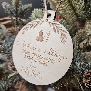 Op de afbeelding: Een ronde, houten kerstbal met een lichtbruine afwerking. De bal heeft gegraveerde tekst: "It takes a village, Thank you for being a part of ours, Love Baby McCue." Een boom en een babyfles zijn ook gegraveerd.