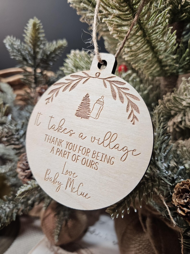 Op de afbeelding: Een ronde, houten kerstbal met een lichtbruine afwerking. De bal heeft gegraveerde tekst: "It takes a village, Thank you for being a part of ours, Love Baby McCue." Een boom en een babyfles zijn ook gegraveerd.