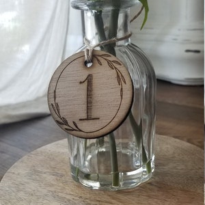 K&ouml;nnte beinhalten: Eine h&ouml;lzerne Tischnummer mit der Nummer "1" eingraviert. Die Tischnummer ist an einer durchsichtigen Glasvase mit einem einzelnen Blumenstiel befestigt.