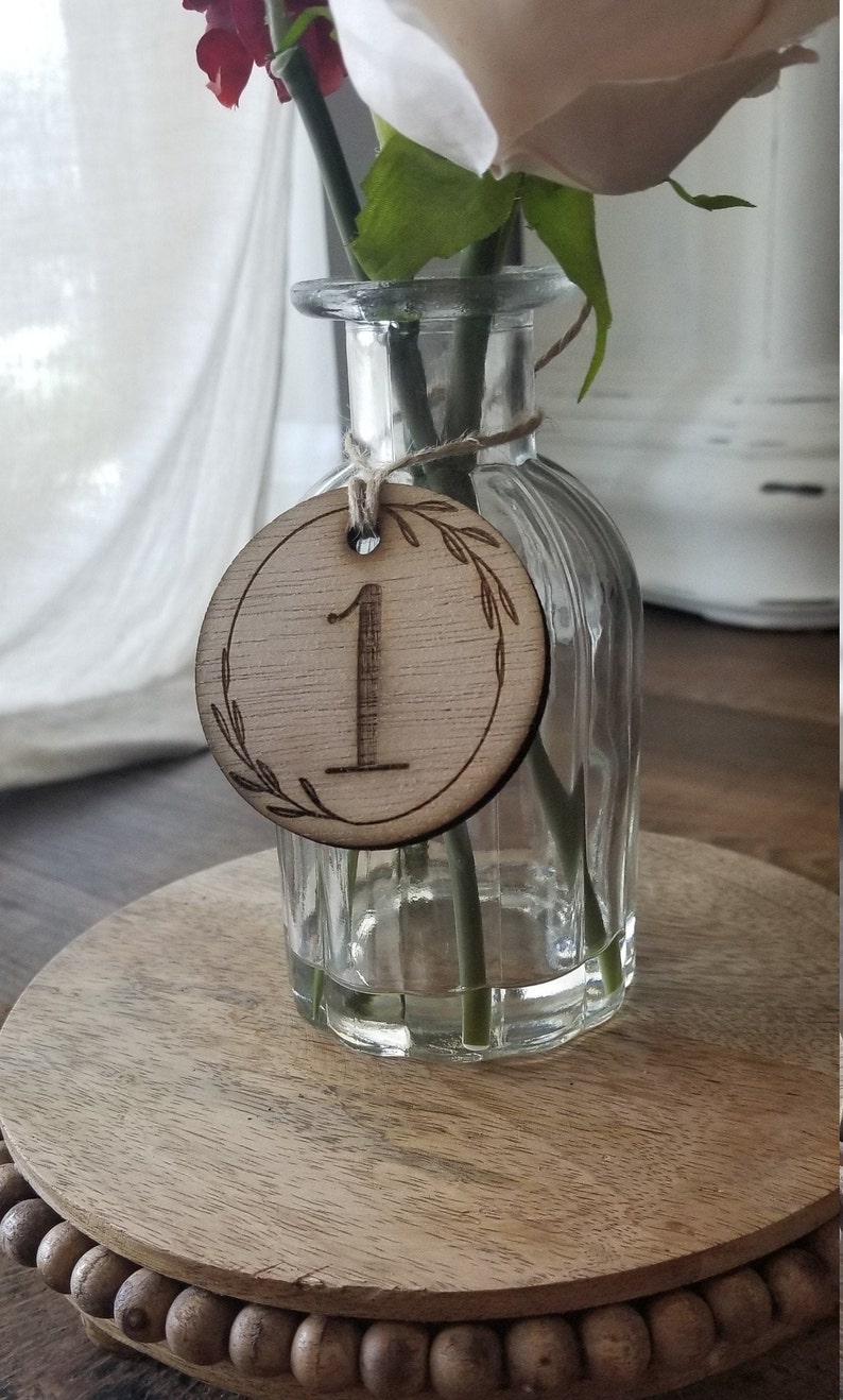 K&ouml;nnte beinhalten: Eine h&ouml;lzerne Tischnummer mit der Nummer "1" eingraviert. Die Tischnummer ist an einer durchsichtigen Glasvase mit einem einzelnen Blumenstiel befestigt.