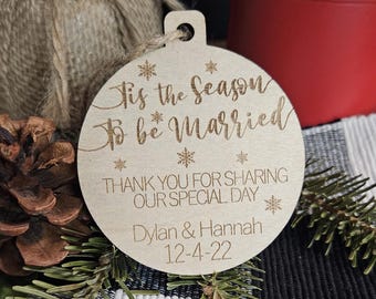 Op de afbeelding: Een houten kerstornament met de tekst "Tis the Season to be Married" en "Thank you for sharing our special day." Bevat de namen Dylan & Hannah en de datum 12-4-22. Het ornament is rond met een gat om op te hangen.