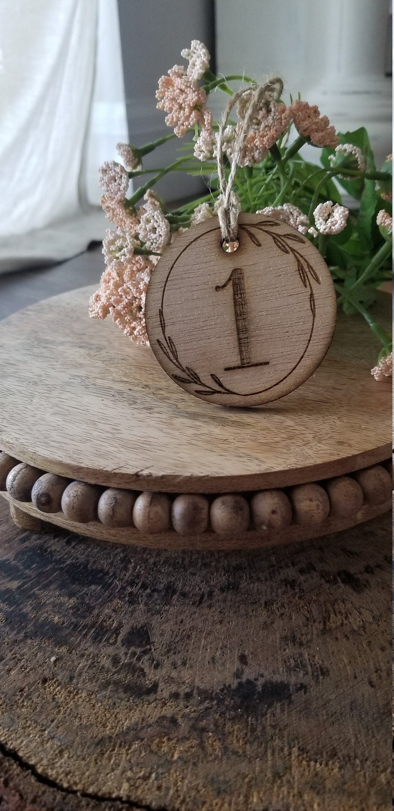 K&ouml;nnte beinhalten: Eine h&ouml;lzerne Tischnummer mit der Nummer "1" eingraviert. Die Nummer ist von einem Kranz aus Bl&auml;ttern umgeben. Die Tischnummer steht auf einem Holzsockel mit einem Perlenrand.