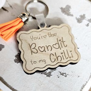 May include: A wooden keychain with the phrase "You're the Bandit to my Chilli" engraved on it. The keychain has a silver ring, a clear plastic clip, and an orange tassel. The wooden piece has a decorative border.