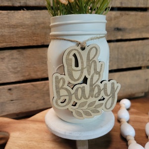 May include: White mason jar with a wooden sign that says "Oh Baby" hanging from it. The jar is filled with a bouquet of small white flowers and is sitting on a white pedestal.