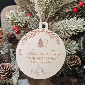Op de afbeelding: Een ronde houten kerstversiering met een boom- en flesontwerp, en de tekst "It takes a village". De versiering hangt aan een bevroren dennentak. De tekst luidt "THANK YOU FOR BEING A PART OF OURS Love Baby McCue".