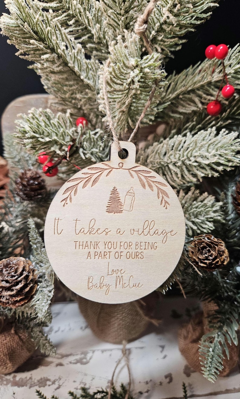 Op de afbeelding: Een ronde houten kerstversiering met een boom- en flesontwerp, en de tekst "It takes a village". De versiering hangt aan een bevroren dennentak. De tekst luidt "THANK YOU FOR BEING A PART OF OURS Love Baby McCue".