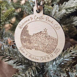 May include: Wooden Christmas ornament with a laser-etched design. The ornament features the text "Welcome to Castle Rock, CO" and a depiction of a local landmark. It also includes the names "Tyler & Ashlee" and the date "Oct 16-17, 2025".