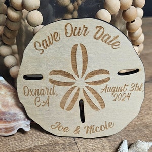 May include: A wooden sand dollar-shaped wedding announcement with the text "Save Our Date", "Oxnard, CA", "August 31st, 2024", and "Joe & Nicole" engraved on it. The announcement is light brown with a dark brown border.