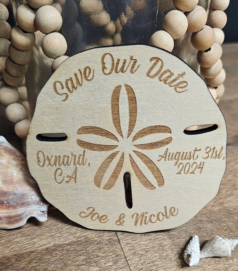 May include: A wooden sand dollar-shaped wedding announcement with the text "Save Our Date", "Oxnard, CA", "August 31st, 2024", and "Joe & Nicole" engraved on it. The announcement is light brown with a dark brown border.