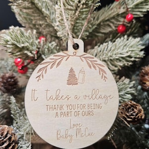 Op de afbeelding: Ronde houten kerstbal met een lichte bruine afwerking. De bal heeft een decoratieve bladerenrand en bevat de tekst "It takes a village" en "Thank you for being a part of ours". Een babyfles en boom zijn ook gegraveerd.