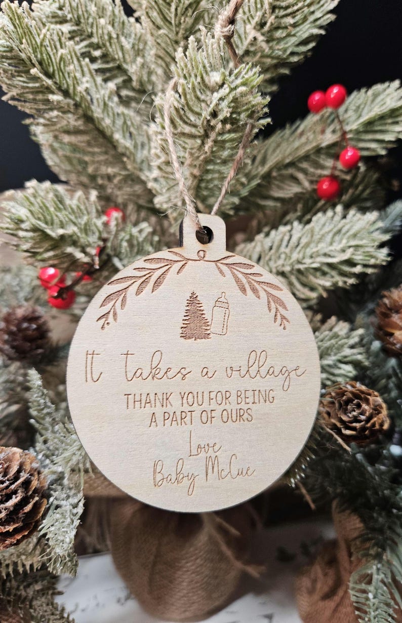 Op de afbeelding: Ronde houten kerstbal met een lichte bruine afwerking. De bal heeft een decoratieve bladerenrand en bevat de tekst "It takes a village" en "Thank you for being a part of ours". Een babyfles en boom zijn ook gegraveerd.
