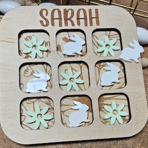 May include: A wooden game board with a tic-tac-toe grid. The board has a light brown finish and features a green floral design. The game is called "Sarah" and includes images of bunnies and flowers.