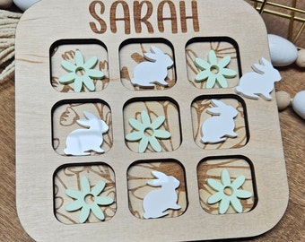 May include: A wooden game board with a noughts and crosses grid. The board has a light brown finish and features a green floral design. The game is called "Sarah" and includes images of bunnies and flowers.