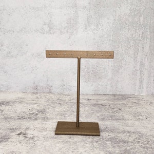 Puede incluir: Soporte de exhibici&oacute;n de aretes de metal dorado con una base rectangular y una barra horizontal con m&uacute;ltiples agujeros para sostener aretes.