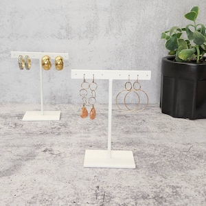 Puede incluir: Expositor de pendientes blanco con tres pares de pendientes dorados. El expositor tiene una base en forma de T y una barra horizontal con varios agujeros para colgar pendientes.