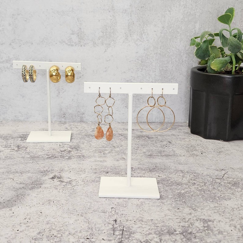 Puede incluir: Expositor de pendientes blanco con tres pares de pendientes dorados. El expositor tiene una base en forma de T y una barra horizontal con varios agujeros para colgar pendientes.