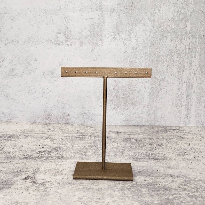 Puede incluir: Soporte de exhibici&oacute;n de aretes de metal dorado con una base rectangular y una barra horizontal con m&uacute;ltiples agujeros para sostener aretes.