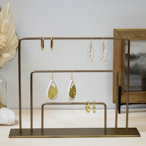 May include: A gold metal earring display stand with three tiers. The stand has a rectangular base and is made of thin metal rods. There are six pairs of earrings on the stand, including hoop earrings, drop earrings with a white background and gold leaf design, and drop earrings with a white background and a green leaf design.