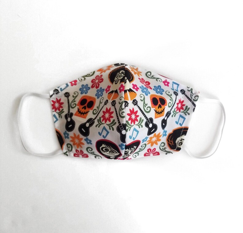 May include: White face mask with a colorful print of skeletons, guitars, and flowers. The print is inspired by the movie Coco.