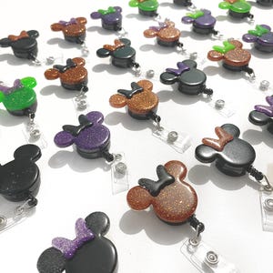 May include: A collection of Minnie Mouse-shaped badge reels in various colors, including black, purple, green, and bronze. Each reel features a glittery bow and a clear plastic clip for attaching to clothing or accessories.