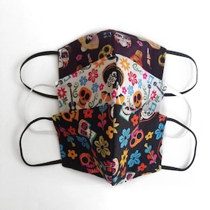 May include: Three colorful face masks with a Day of the Dead theme. The masks feature skulls, flowers, and musical notes. The masks are made of fabric and have elastic ear loops.