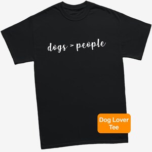 May include: Black t-shirt with the white text "dogs > people". An orange rectangle in the bottom right corner reads "Dog Lover Tee". The shirt is a casual wear item.