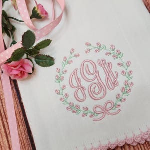 May include: White embroidered cloth with a pink floral wreath and monogram. The monogram features the letters "GJ" in a delicate script. A pink ribbon and roses are placed near the cloth, with a scalloped pink trim.