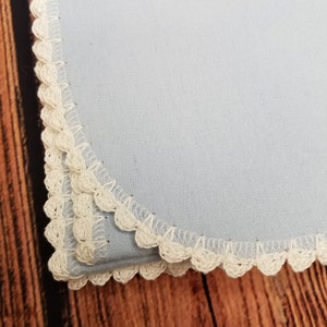 May include: A light blue cloth napkin with a decorative, off-white crocheted trim. The napkin is square-shaped and appears to be made of a soft fabric. The crocheted trim adds a delicate, handmade touch to the napkin's edge.