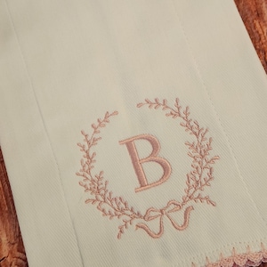 May include: White cloth with a pink embroidered wreath design, featuring the letter "B" and a bow. The edge is trimmed with a pink lace border. The background is a wooden surface.