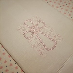 May include: White cloth with a pink embroidered cross design. The cross features a floral center and decorative swirls. The cloth is folded, with a detailed hem visible. The background has a floral pattern.