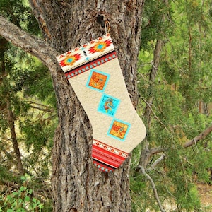 May include: A beige Christmas stocking with a Southwestern design hangs from a tree. The stocking features colorful geometric patterns in red, orange, turquoise, and black. The design includes three turquoise squares with a decorative pattern.