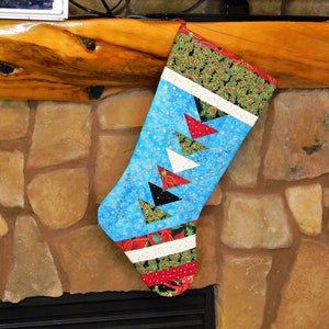 May include: A quilted Christmas stocking with a blue background and colorful triangle accents. The stocking features a green and red holly berry pattern at the top and bottom. The stocking is hanging on a wooden mantle.