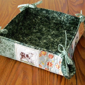 May include: A handmade fabric basket with a farm theme. The interior is a dark green leaf pattern, while the exterior features a patchwork design with a cow and floral accents. The corners are tied with green bows.