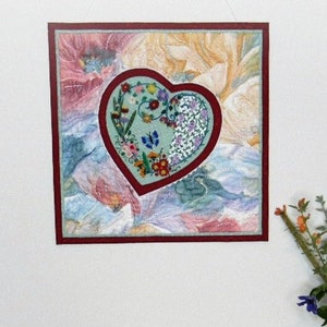 May include: A framed embroidery of a heart with floral designs in shades of green, blue, and red, hanging on a white wall above a wooden table with a vase of flowers.