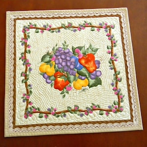 May include: A square quilted placemat featuring a central image of a colorful fruit arrangement, including grapes, pears, and peppers. The design is framed by a brown border and a delicate lace edge. The background has a quilted pattern.