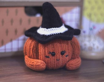 May include: A knitted orange pumpkin plush toy featuring a black witch hat with a white band. The pumpkin has two black bead eyes, stitched eyelashes, and a small embroidered mouth. The texture is ribbed, and the pumpkin has two small arms.
