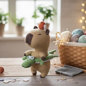 May include: A knitted capybara toy wearing a green alligator floatie, with a small bird on its head. A basket of yarn balls, knitting needles, and buttons are on a wooden surface. The scene is illuminated by fairy lights.