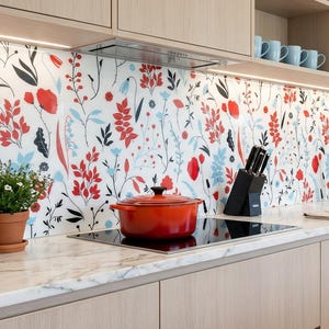 May include: A kitchen featuring light wood cabinets, a white marble worktop, and a splashback with a colourful floral pattern of red, blue, and black botanical illustrations. A bright orange pot sits on the hob next to a black knife block.