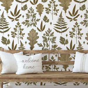 Può includere: Un muro bianco con un motivo di carta da parati botanica marrone e verde. Una panca di legno con cuscini bianchi e un cuscino con il testo "welcome home" è davanti al muro. Una pianta verde in vaso è sul lato destro dell'immagine.