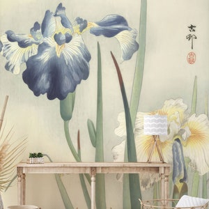 May include: A vintage-style wallpaper mural featuring a close-up of blue and white iris flowers with green leaves. The wallpaper is displayed in a living room setting with a wooden console table, wicker baskets, and a throw pillow.