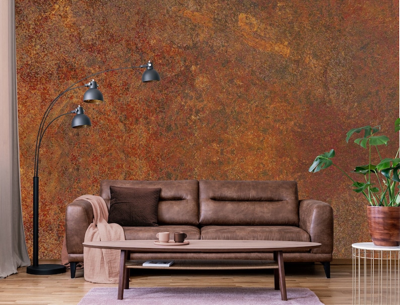 May include: A brown leather sofa with a throw blanket and a brown pillow sits in front of a wall with a rusty orange and brown textured wallpaper. A floor lamp with three black shades stands to the left of the sofa. A potted plant with green leaves sits on a gold metal side table to the right of the sofa.