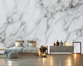 Puede incluir: Un interior de dormitorio con una pared con estampado de mármol blanco, una cama con ropa de cama y almohadas blancas, un suelo de madera, una mesita de noche con una lámpara, una cómoda con jarrones negros y velas, y un cuadro enmarcado en el suelo.
