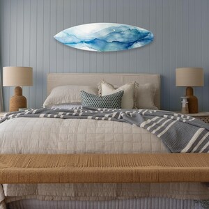 May include: A bedroom scene featuring a bed with a beige comforter and a gray and white striped blanket. Above the bed is a surfboard-shaped wall art with blue and white watercolor designs. A woven bench sits at the foot of the bed.