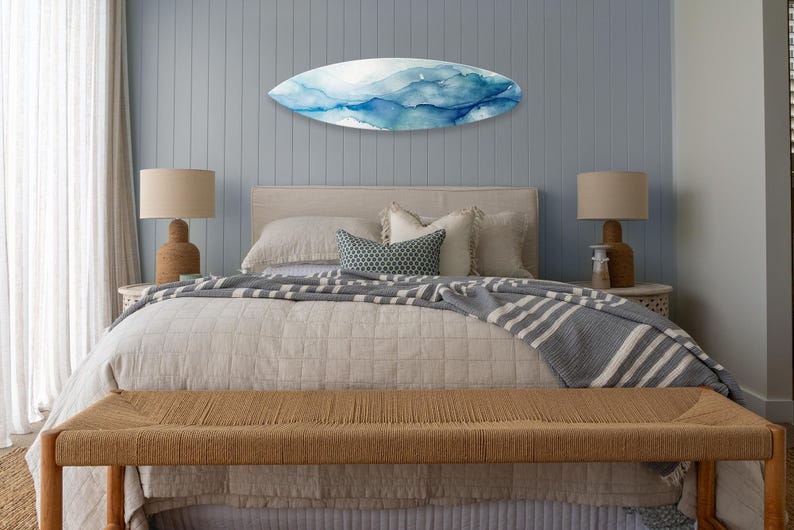 May include: A bedroom scene featuring a bed with a beige comforter and a gray and white striped blanket. Above the bed is a surfboard-shaped wall art with blue and white watercolor designs. A woven bench sits at the foot of the bed.