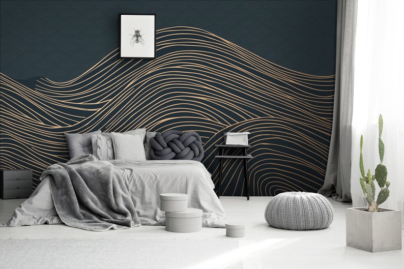 May include: A bedroom with a dark blue wall featuring a gold and black abstract wave pattern wallpaper. A gray bed with a gray blanket and pillows is in front of the wall. A gray pouf sits on the floor in the foreground.