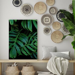 Puede incluir: Una pared blanca con una colección de cestas de pared tejidas en varios tamaños y patrones. Una gran impresión de lienzo de hojas tropicales verdes cuelga sobre un banco de madera con una manta blanca y dos cestas tejidas.