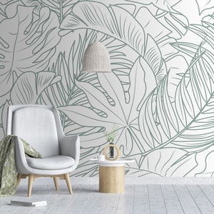 Puede incluir: Un sill&oacute;n blanco con una manta de tiro verde claro se encuentra frente a una pared con un patr&oacute;n de hojas tropicales blancas y verdes.