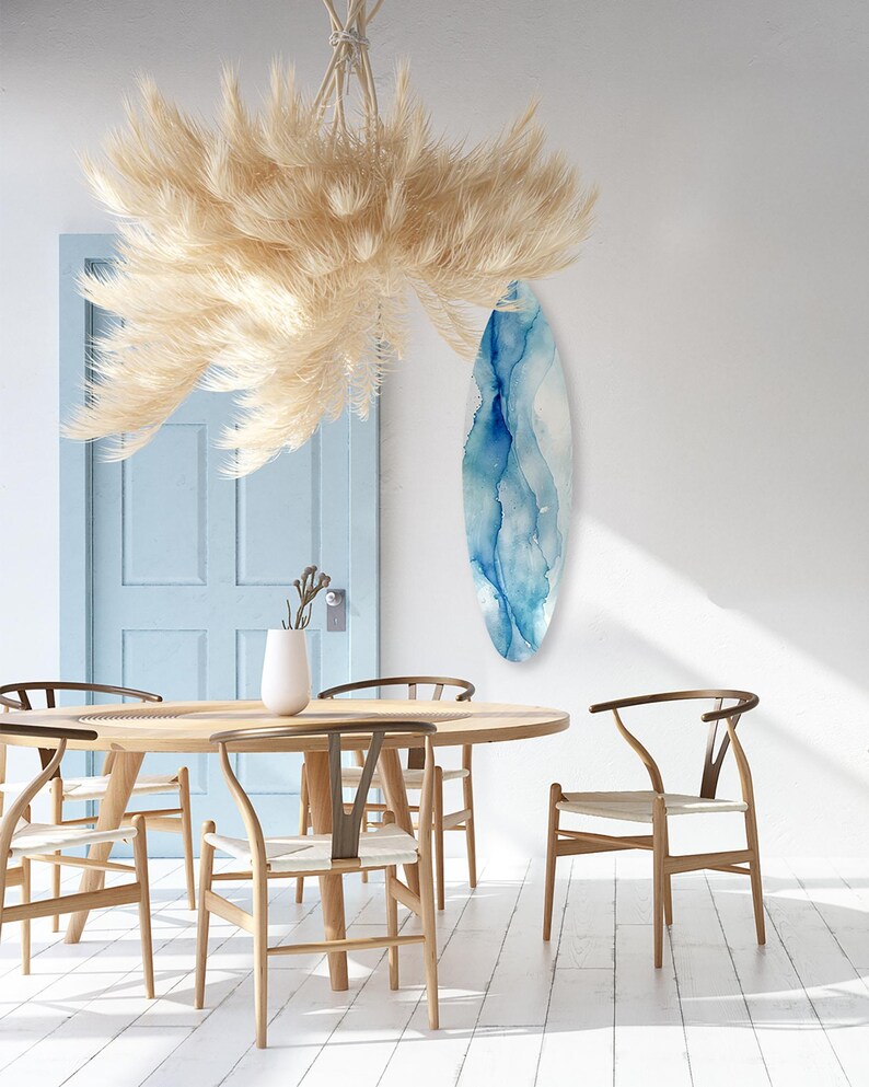 May include: A dining room scene featuring a light wood round table and six matching chairs with woven seats. A large, feathery, beige chandelier hangs above the table. A blue and white abstract art piece hangs on the white wall.